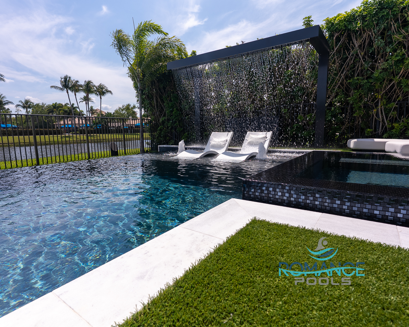 Sun shelf pool loungers under waterfall feature with palm trees and modern landscape design by Romance Pools